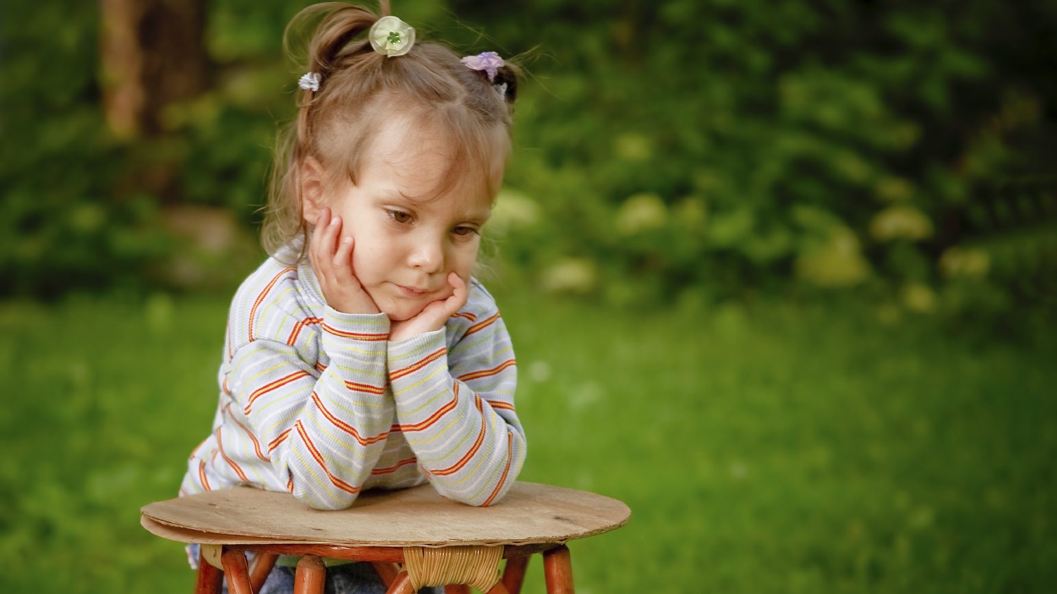 Little girl on a green lawn leans against chair and thinks of mother.
