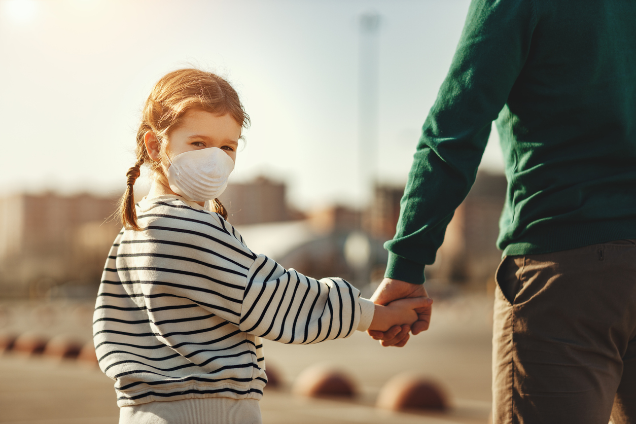 Back view of little girl in medical mask looking at camera over shoulder and holding hand of crop father while walking on parking lot during coronavirus outbreak