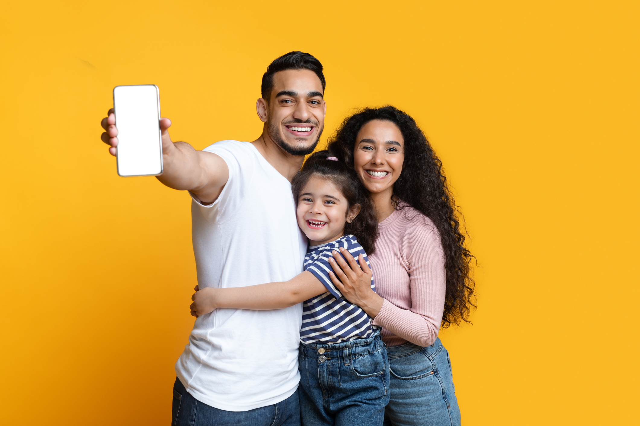 Cheerful Arab Family Of Three Demonstrating Blank Smartphone With White Screen, Loving Middle Eastern Mom, Dad And Little Daughter Embracing And Enjoying New Mobile Application Or Website, Mockup